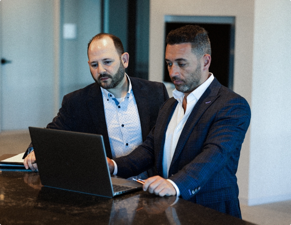 2 men in suite looking at laptop on dark marble counter