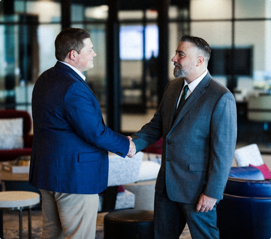 2 men in dark suits shaking hands staring at each other greeting in lobby with couches